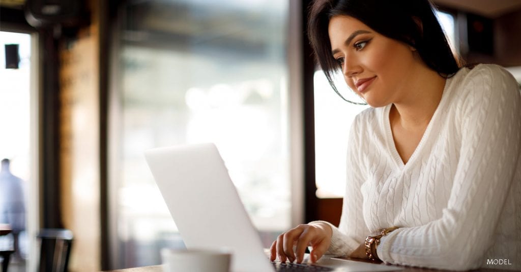 Dark-haired young woman in a white sweater looking at her laptop screen