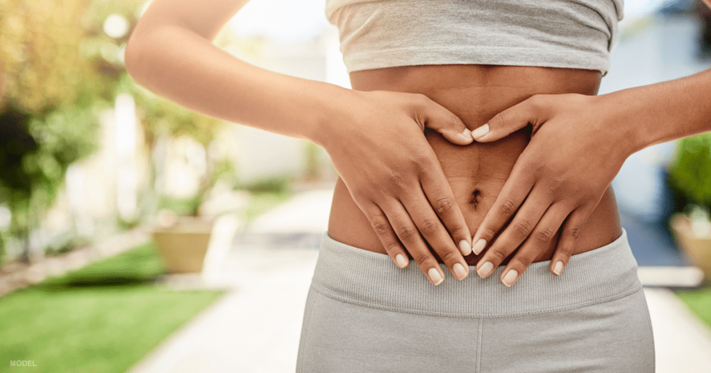 Abdomen of fit woman outdoors with grey tank top pulled up to expose tummy and hands forming a heart shape over her belly button