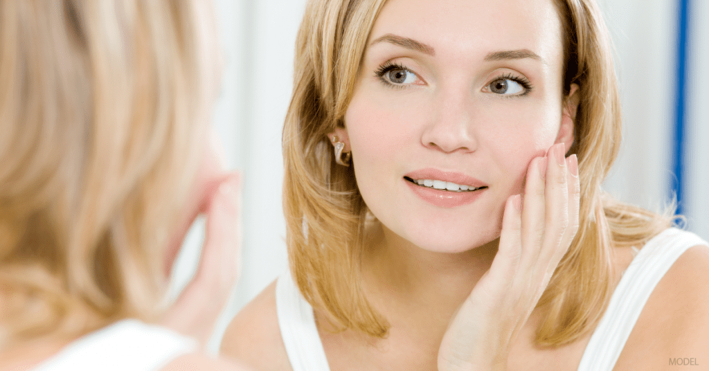 Face of woman with blonde hair and brown eyes looking in the mirror while touching her cheek