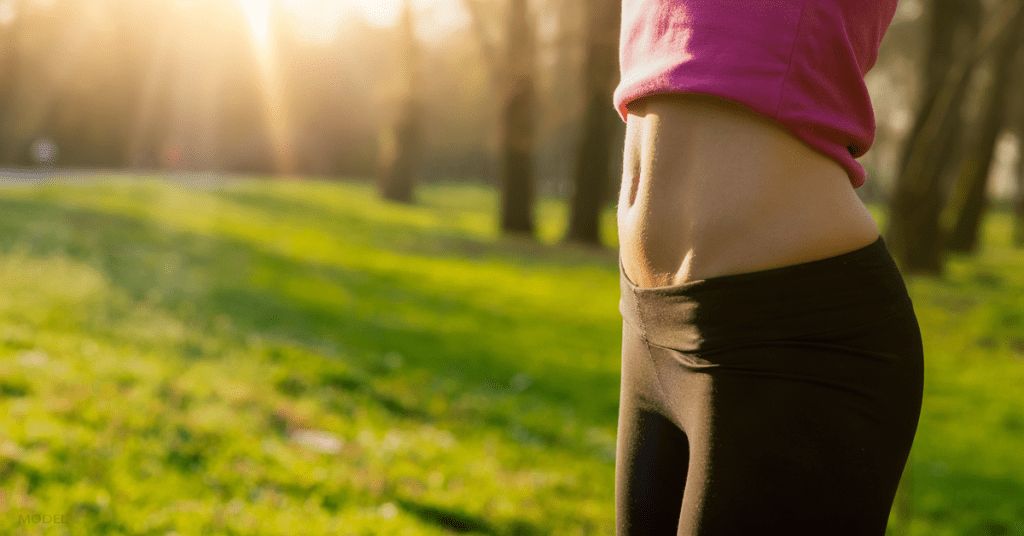 Torso of a woman wearing black yoga pants standing in grass with toned abdomen exposed