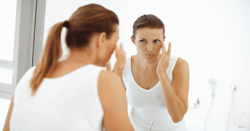 Woman with long brown hair in a ponytail looking in the mirror and touching her cheek