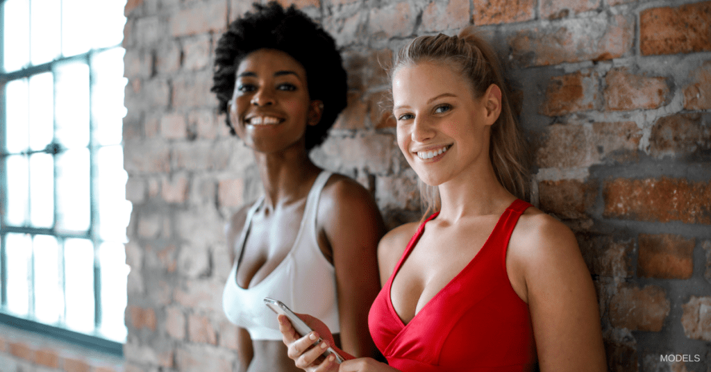 Two women standing against brick wall smiling. (Models)
