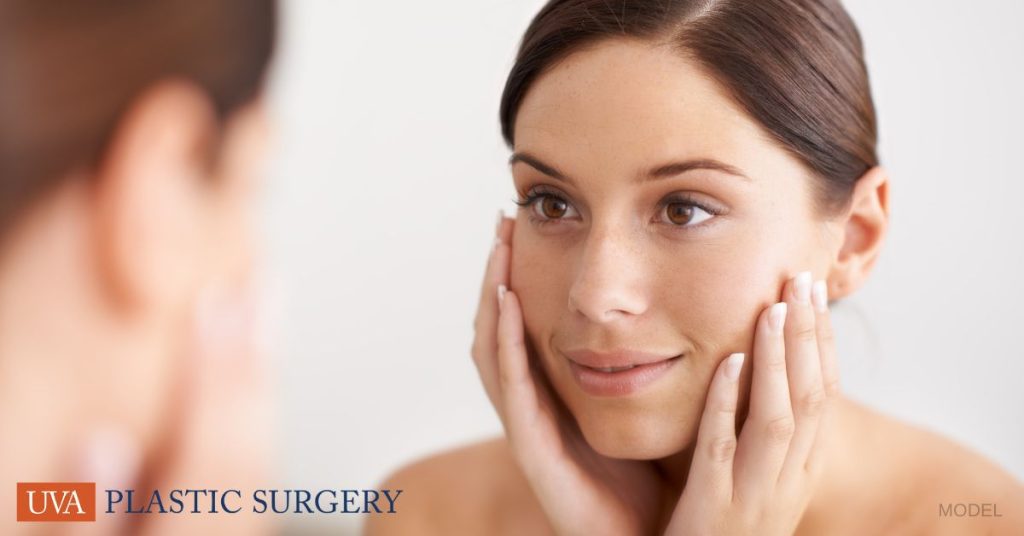 Woman holding face and smiling while looking in the mirror. (Model)